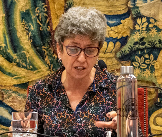 Retrato de Ángeles Ezama hablando en una conferencia. Mujer de cabellos cortos y ondulados, canos. Lleva gafas de montura oscura y pendientes de aro plateados. Detrás de ella, hay un tapiz de colores amarillos, azules y verdes que representa una escena paisajística. Delante de ella, un vaso de agua y una botella.