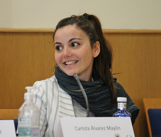 Retrato de Carlota Álvarez Maylín. Mujer joven, delgada, de cabello castaño recogido en una coleta alta. Sonríe mostrando la dentadura y con la mirada hacia la izquierda. Está en una mesa de conferencias, con un micrófono delante.